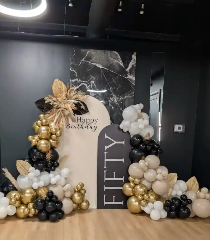 Sophistication defines this stunning fiftieth birthday celebration backdrop, designed specifically to capture memories. A cascading balloon garland featuring metallic gold, matte black, crisp white, and soft nude tones frames the setup elegantly. Textural elements like dried palm spears and fluffy pampas grass intermingle with the globes, adding a modern, organic flair to the arrangement that feels very current.
Centrally positioned are two distinct arch-shaped panels creating depth against a dark wall that features a striking marble insert. The lighter beige arch reads "Happy Birthday" in a delicate script, while a bold black panel prominently displays "FIFTY" in large, vertical block letters. This specific configuration provides an excellent focal point where guests can gather to take memorable pictures throughout the night.
Executing refined 50th birthday party ideas often involves creating a dedicated, stylish space for photography just like this example. The clever combination of classic color palettes and trendy textures offers a mature yet festive atmosphere suitable for marking half a century. It successfully transforms a simple room corner into a major statement piece honoring the birthday person.